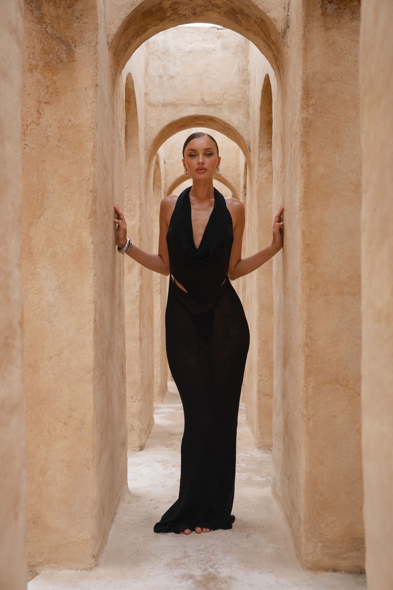 Model wearing a black halter neck dress standing in a sunlit archway with sand-coloured walls.