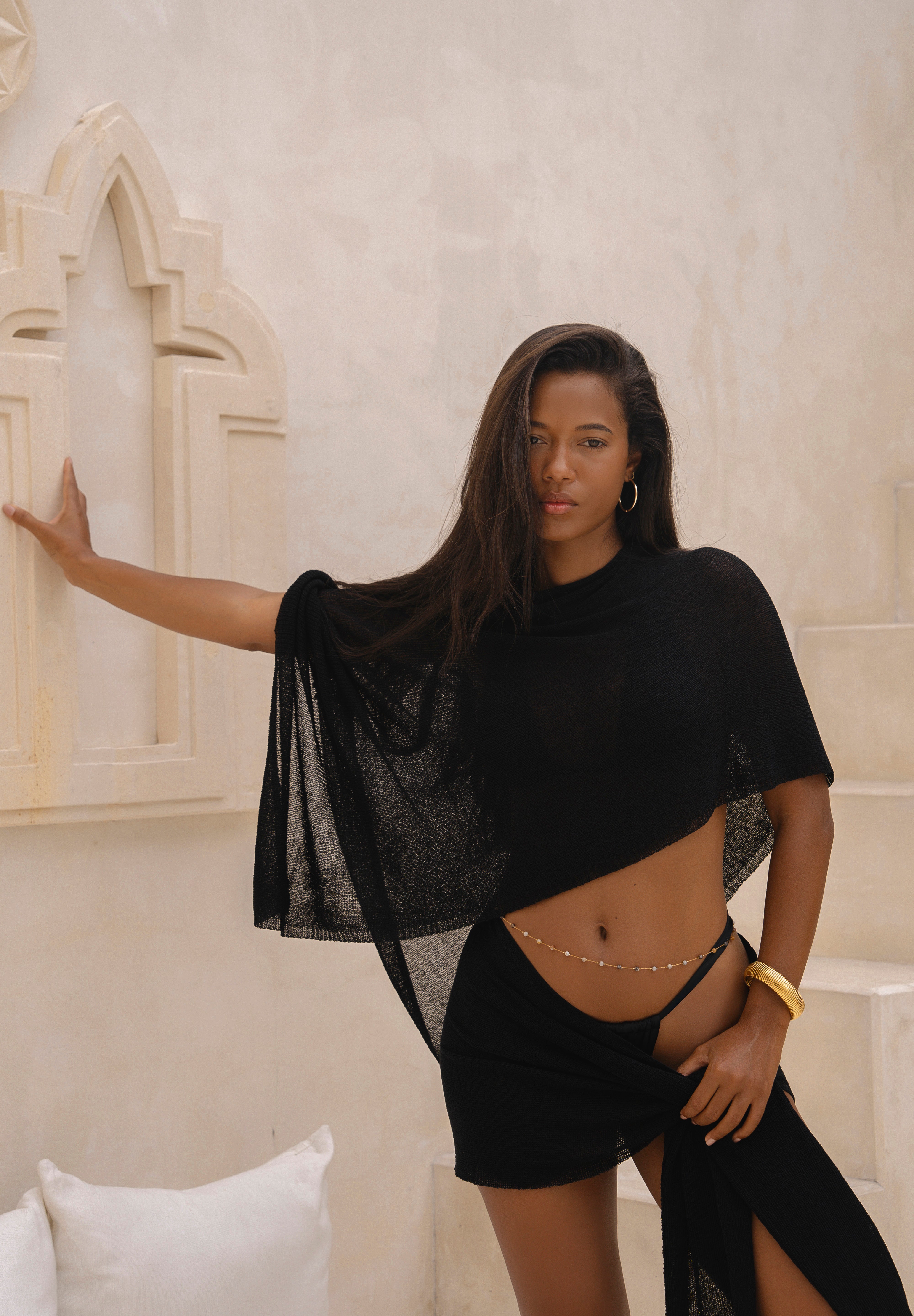 Model wearing a black sarong and black matching cape, posing in front of a neutral background