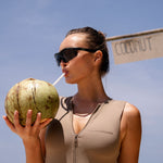 Close up model wearing a cinnamon coloured zip up one piece swimsuit holding a coconut on a beach with a clear blue sky.