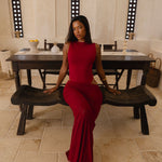 Model wearing a cherry Rogue Season one-piece swimsuit with side cut-out and matching sheer skirt, seated indoors in a neutral setting, sitting on a wooden bench in a room with a large mirror.