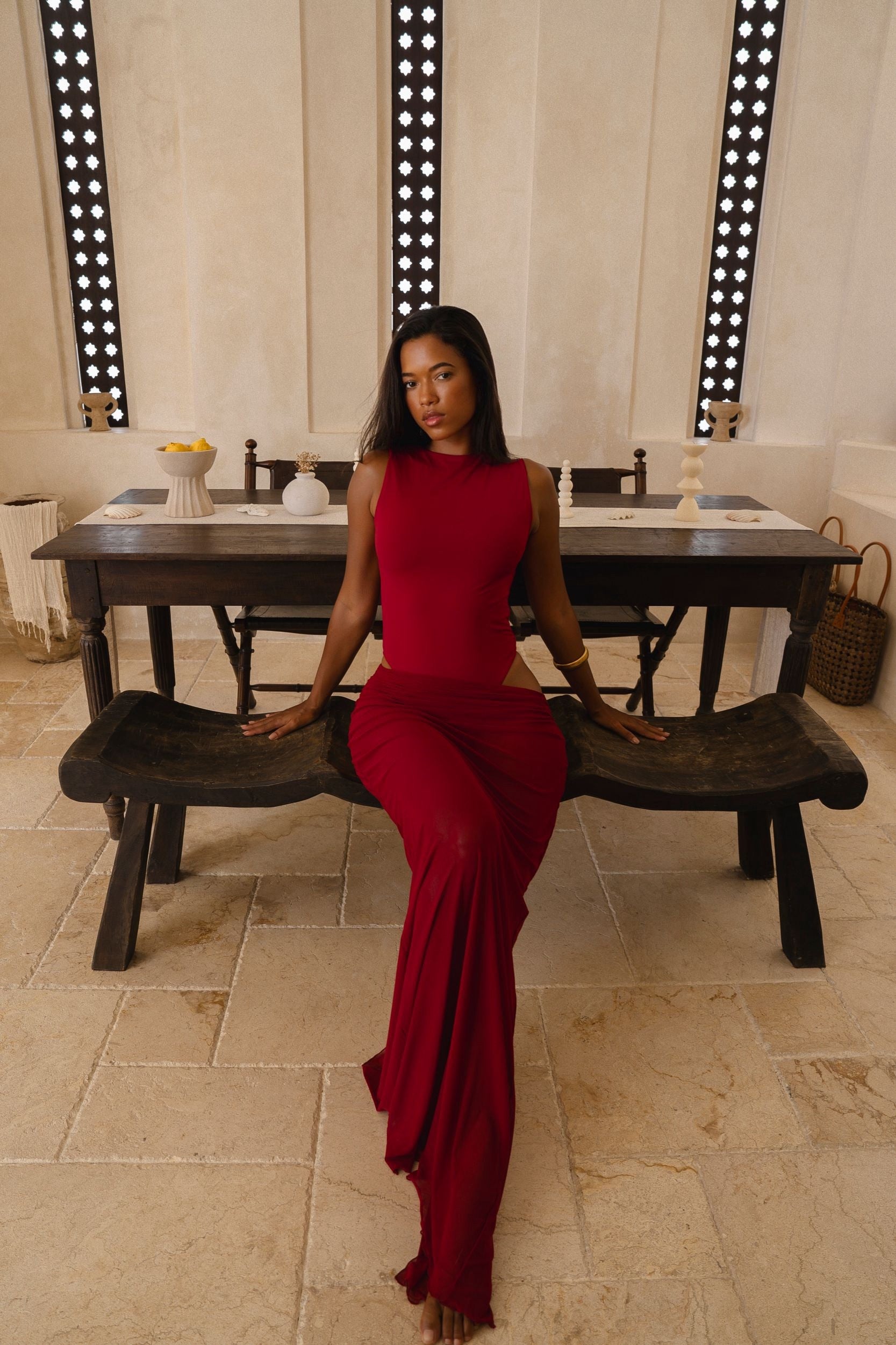Model wearing a cherry Rogue Season one-piece swimsuit with side cut-out and matching sheer skirt, seated indoors in a neutral setting, sitting on a wooden bench in a room with a large mirror.