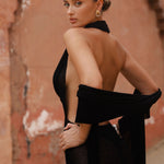 Back shot of model wearing a black halter neck maxi dress standing in a sunlit archway with sand-coloured walls.