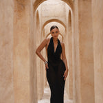 Model wearing a black halter neck dress standing in a sunlit archway with sand-coloured walls.