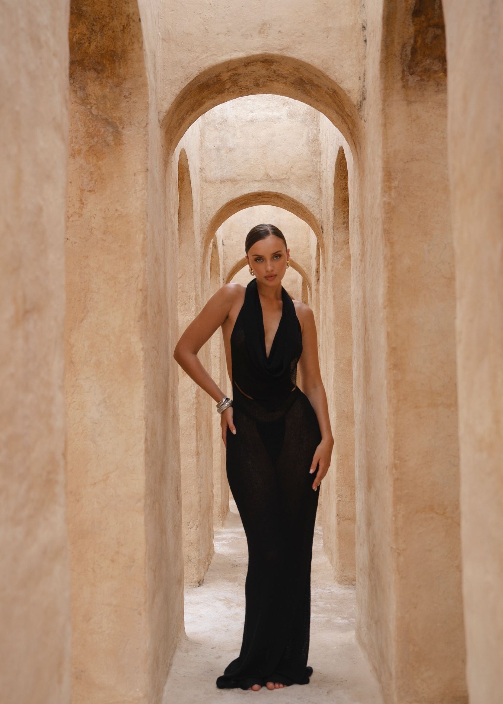 Model wearing a black halter neck dress standing in a sunlit archway with sand-coloured walls.