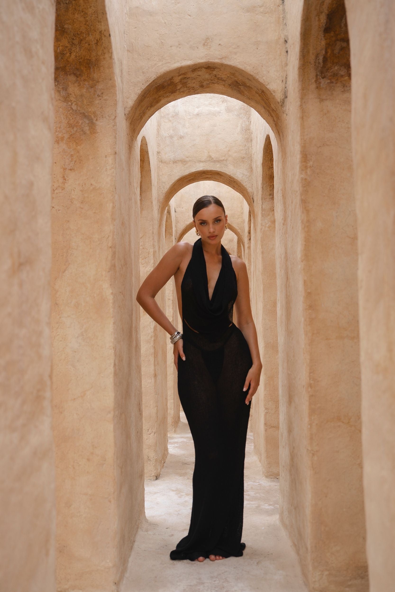 Model wearing a black halter neck dress standing in a sunlit archway with sand-coloured walls.
