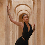 Model wearing a black halter neck dress standing in a sunlit archway with sand-coloured walls.
