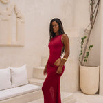Model wearing a cherry Rogue Season one-piece swimsuit with side cut-out and matching sheer skirt, standing in a modern indoor setting with beige walls and decor.