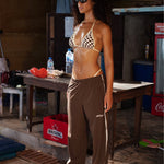 Model wearing a brown and pastel yellow checkered triangle bikini top with brown track pants featuring cream side stripes, brown sneakers, and black sunglasses, standing outdoors in a rustic tropical setting.