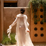 Back shot of model wearing an ivory sheer ruffled maxi cover-up with an ivory bikini, standing outdoors in sunlight against a textured wall with greenery.
