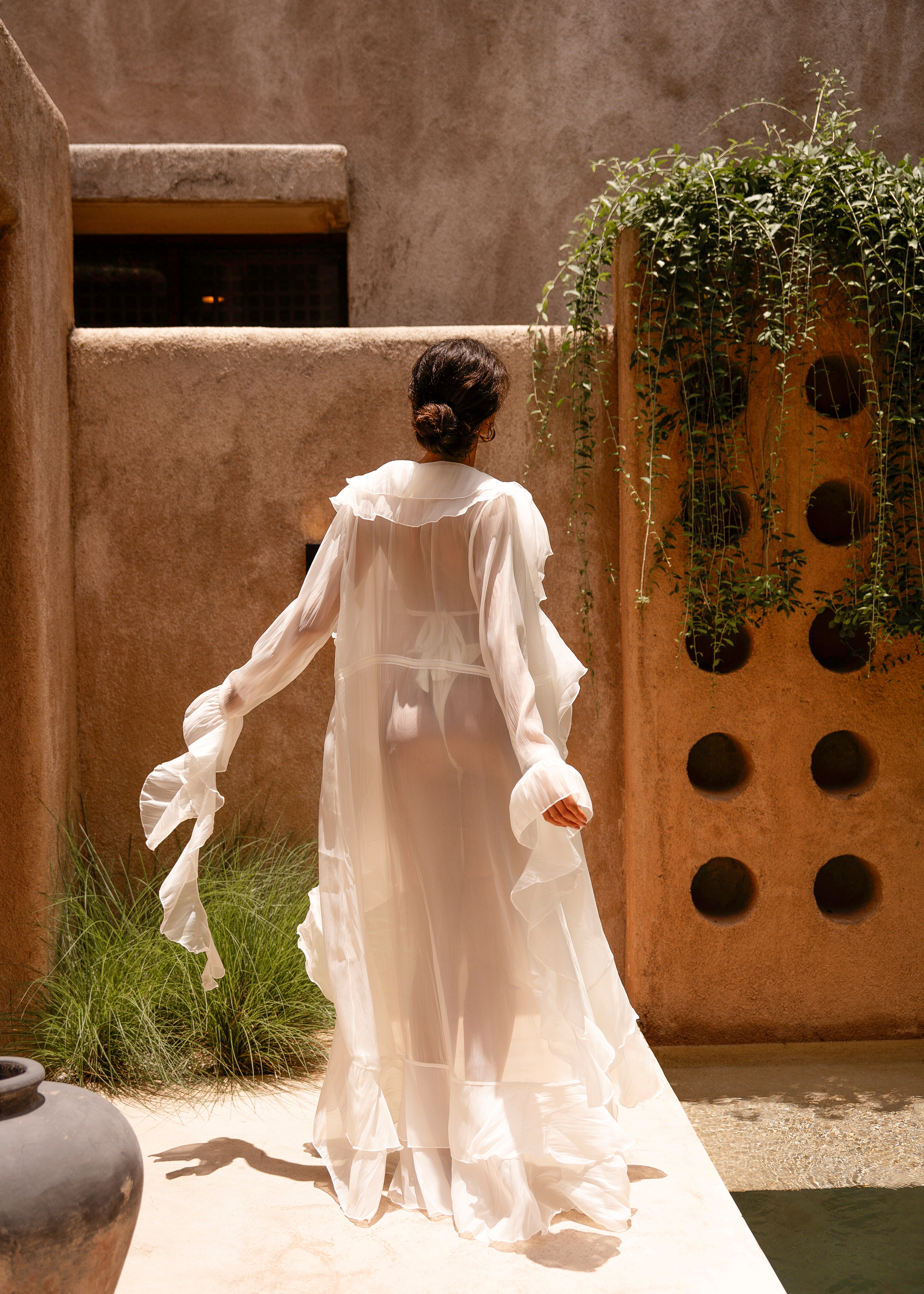 Back shot of model wearing an ivory sheer ruffled maxi cover-up with an ivory bikini, standing outdoors in sunlight against a textured wall with greenery.