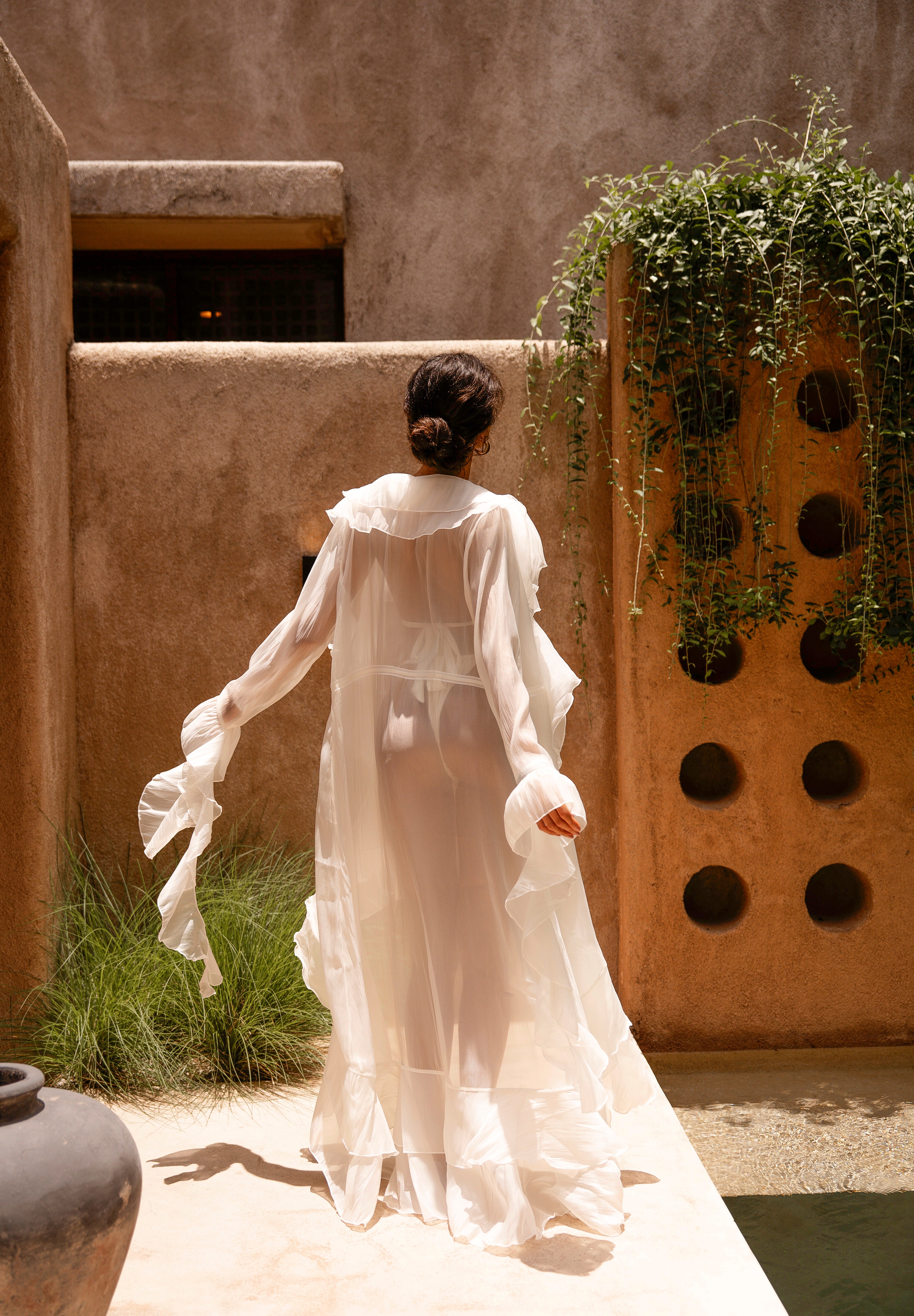 Back shot of model wearing an ivory sheer ruffled maxi cover-up with an ivory bikini, standing outdoors in sunlight against a textured wall with greenery.
