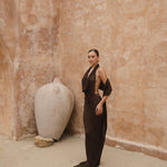 Model in a brown maxi halter dress standing against a textured beige wall with a large white vase.