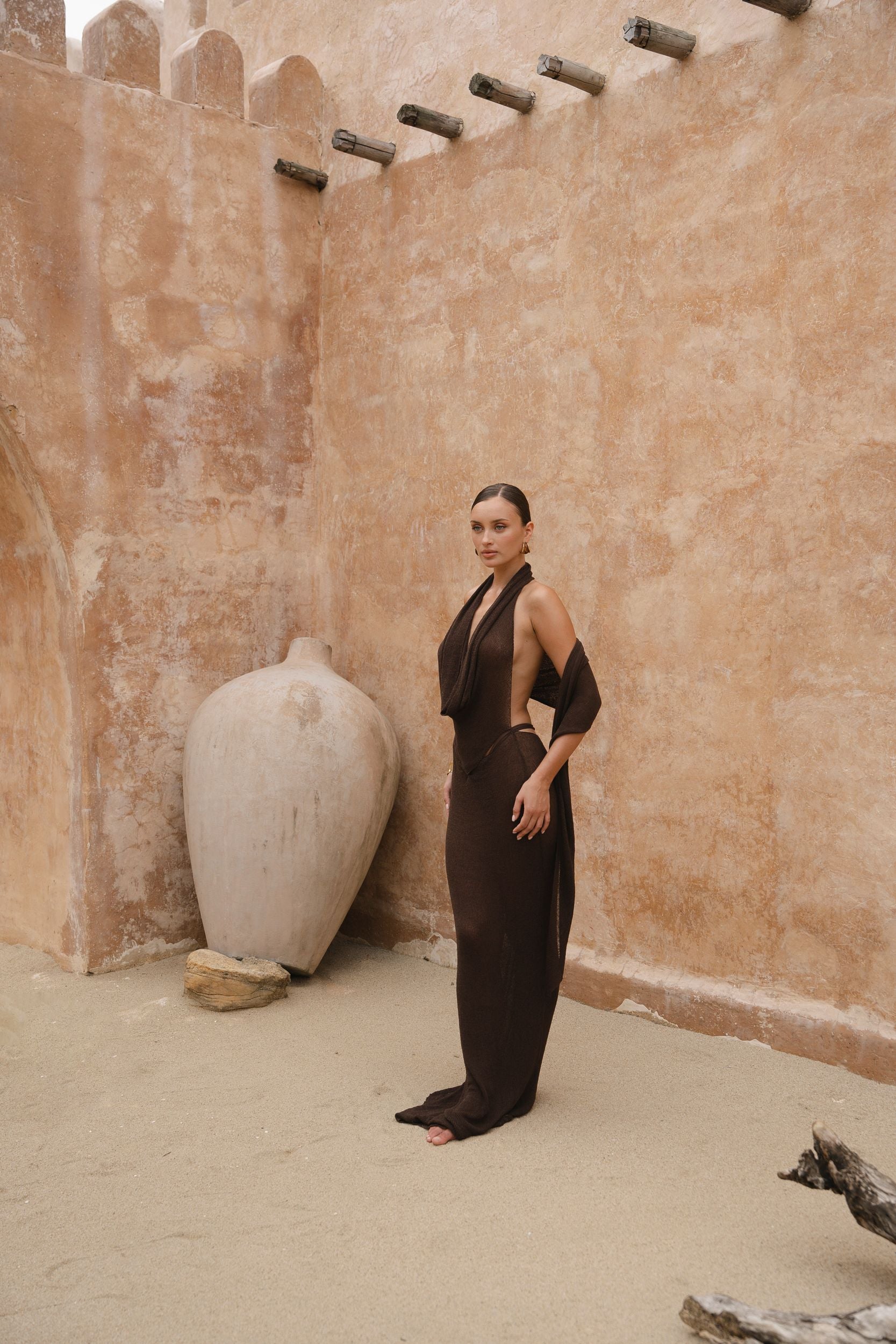 Model in a brown maxi halter dress standing against a textured beige wall with a large white vase.
