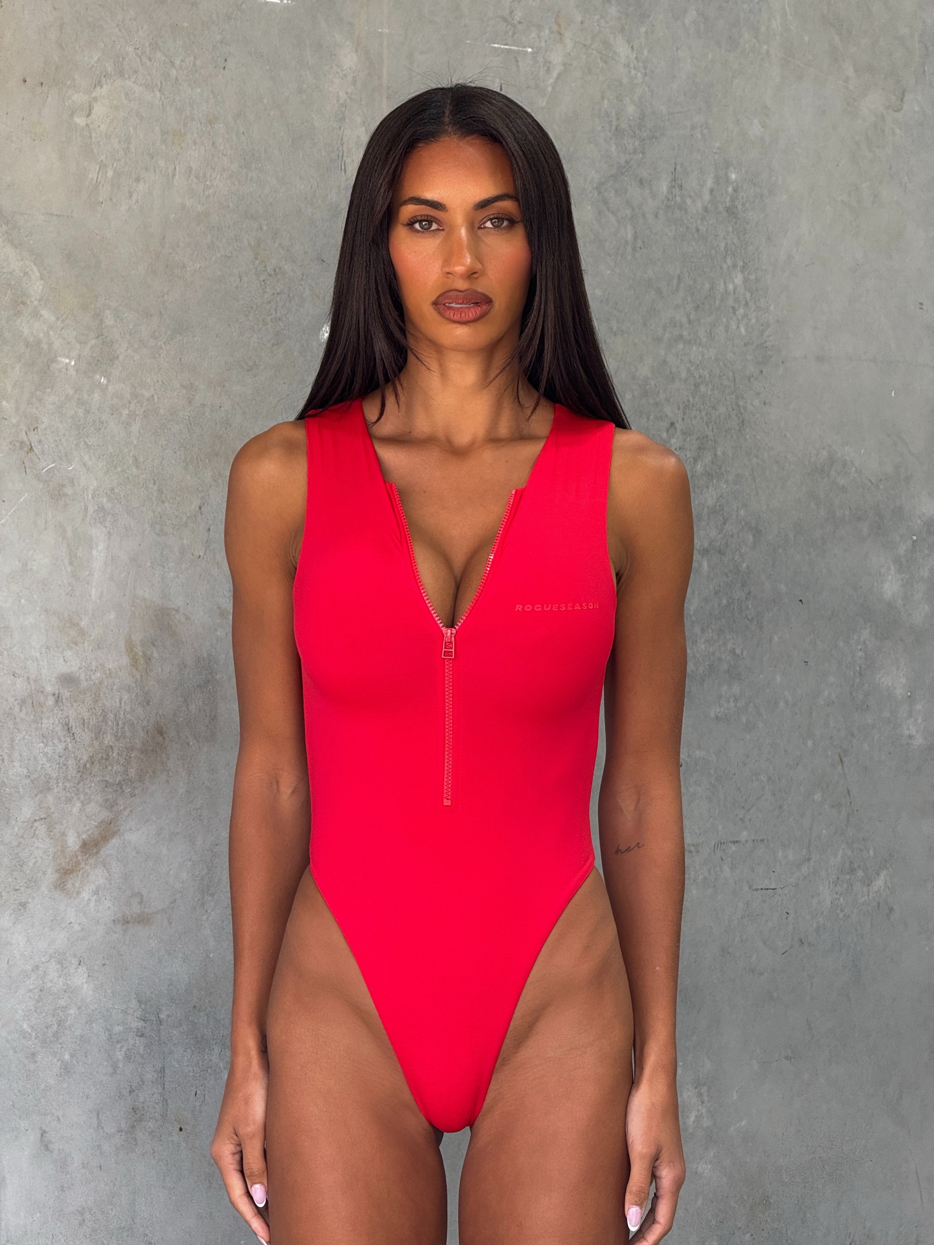 Close up shot of model in a red one piece zip up swimsuit  standing in front of a grey wall.