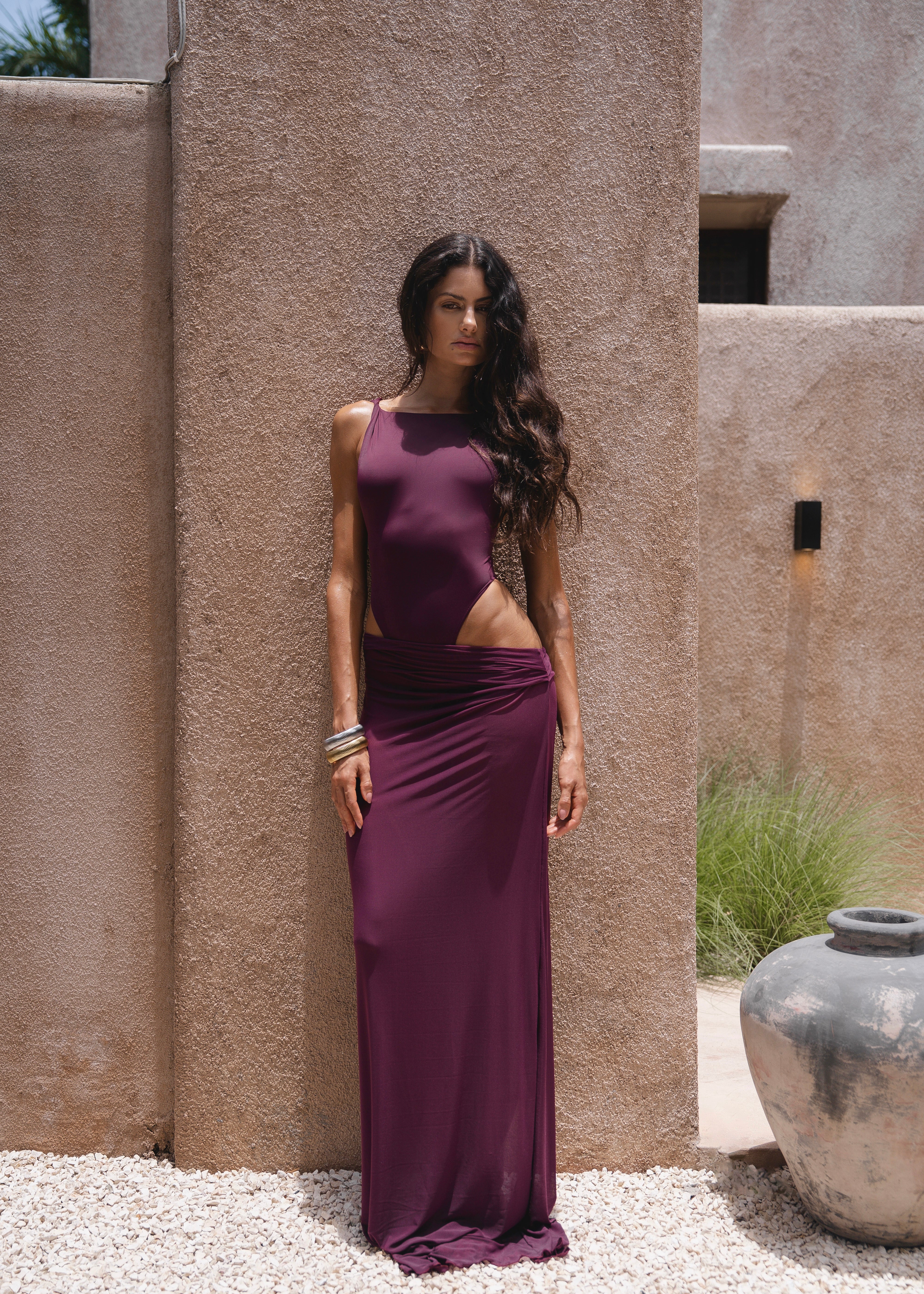 Model wearing a burgundy swimsuit and matching sheer maxi skirt near a textured wall