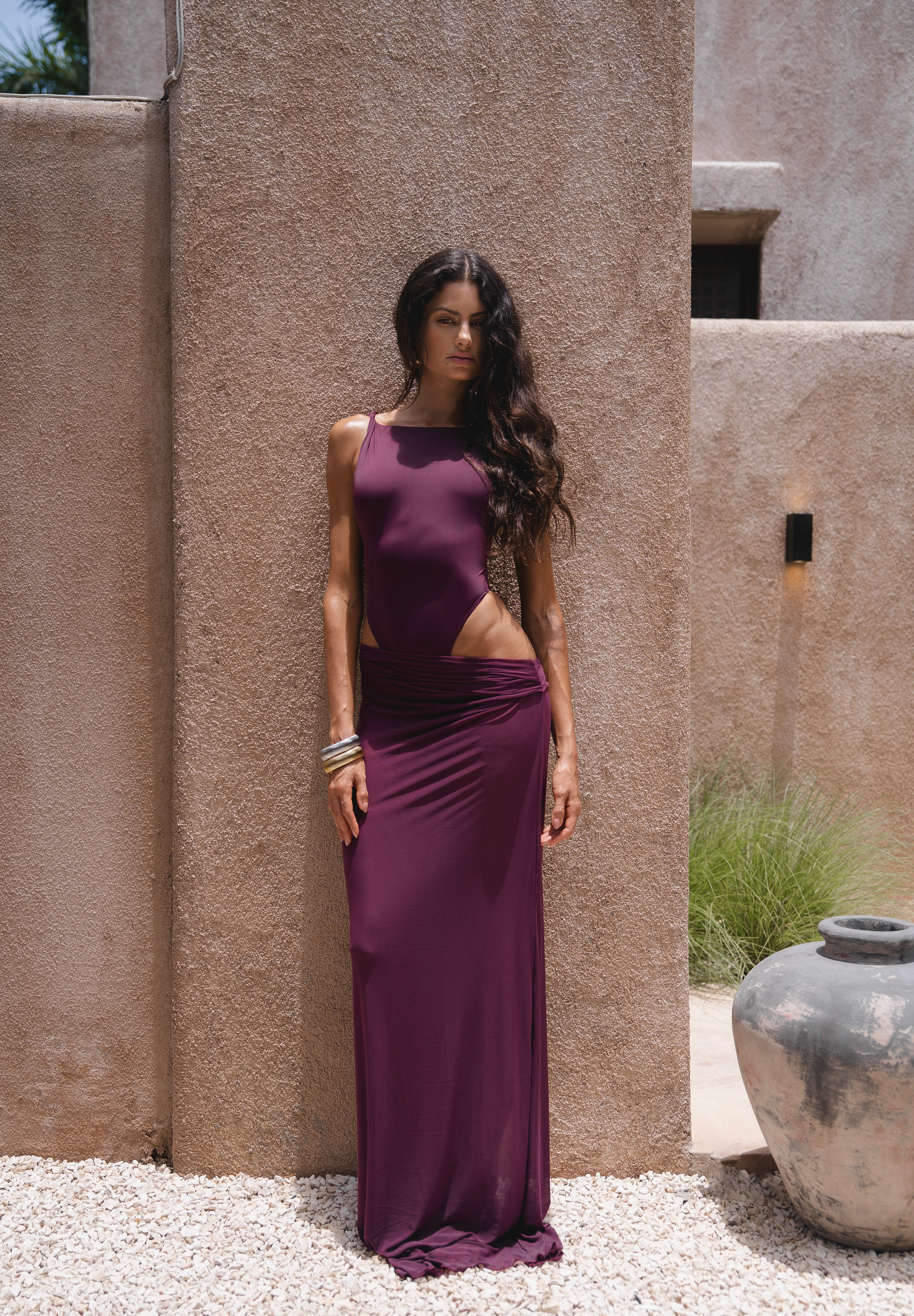 Model wearing a burgundy swimsuit and matching sheer maxi skirt near a textured wall