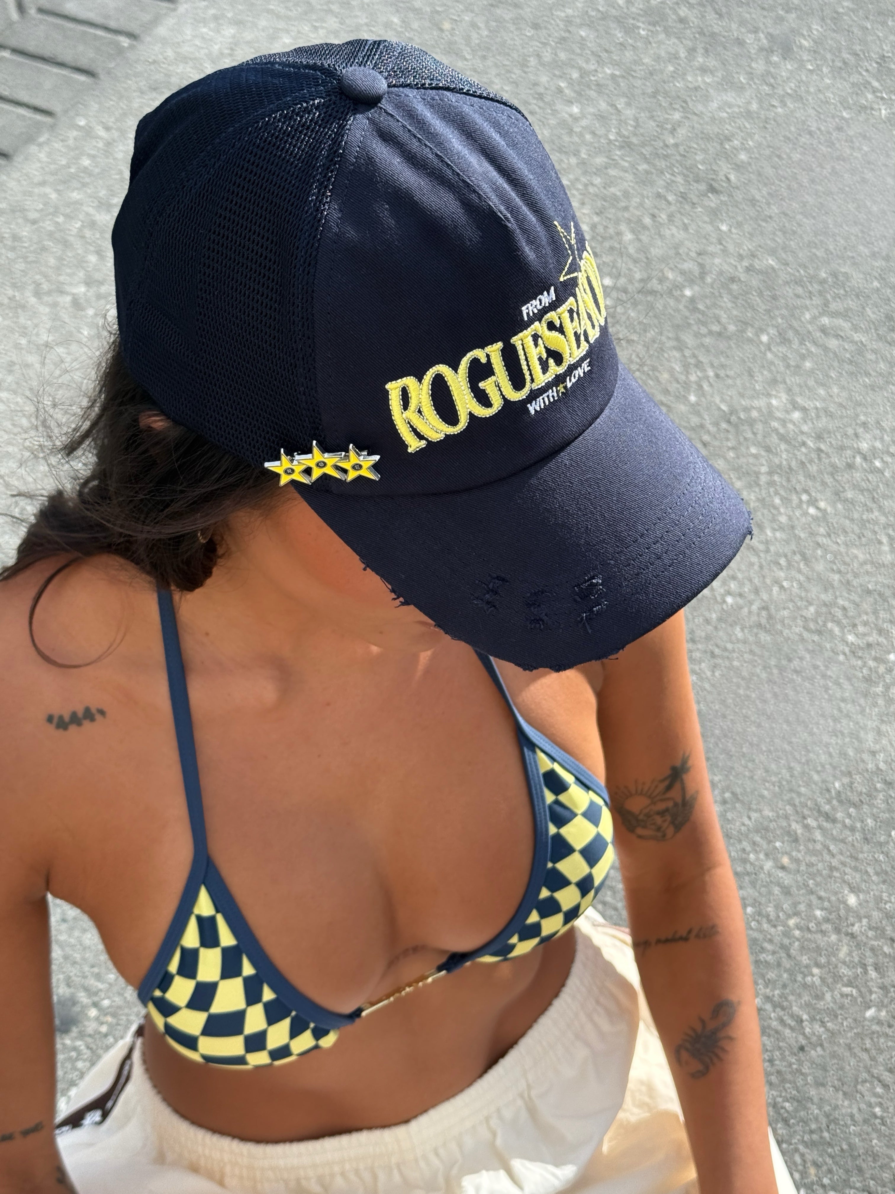Model wearing a checkered bikini in Navy and Yellow and 'ROGUESEASON' branded cap standing on a street 