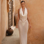 Model wearing an Ivory halter neck maxi dress standing in a sunlit archway with sand-coloured walls.
