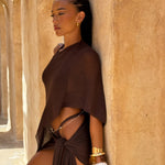 Side shot of model wearing a brown cape and sarong with matching bikini bottom, styled back with  gold and white jewellery in front a beige background.