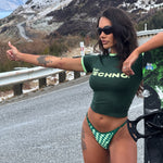 Model wearing black sunglasses, a green Rogue Season cropped t-shirt with printed text 'PLAY TECHNO' and green checkered bikini bottoms, standing on a road in a snowy setting.