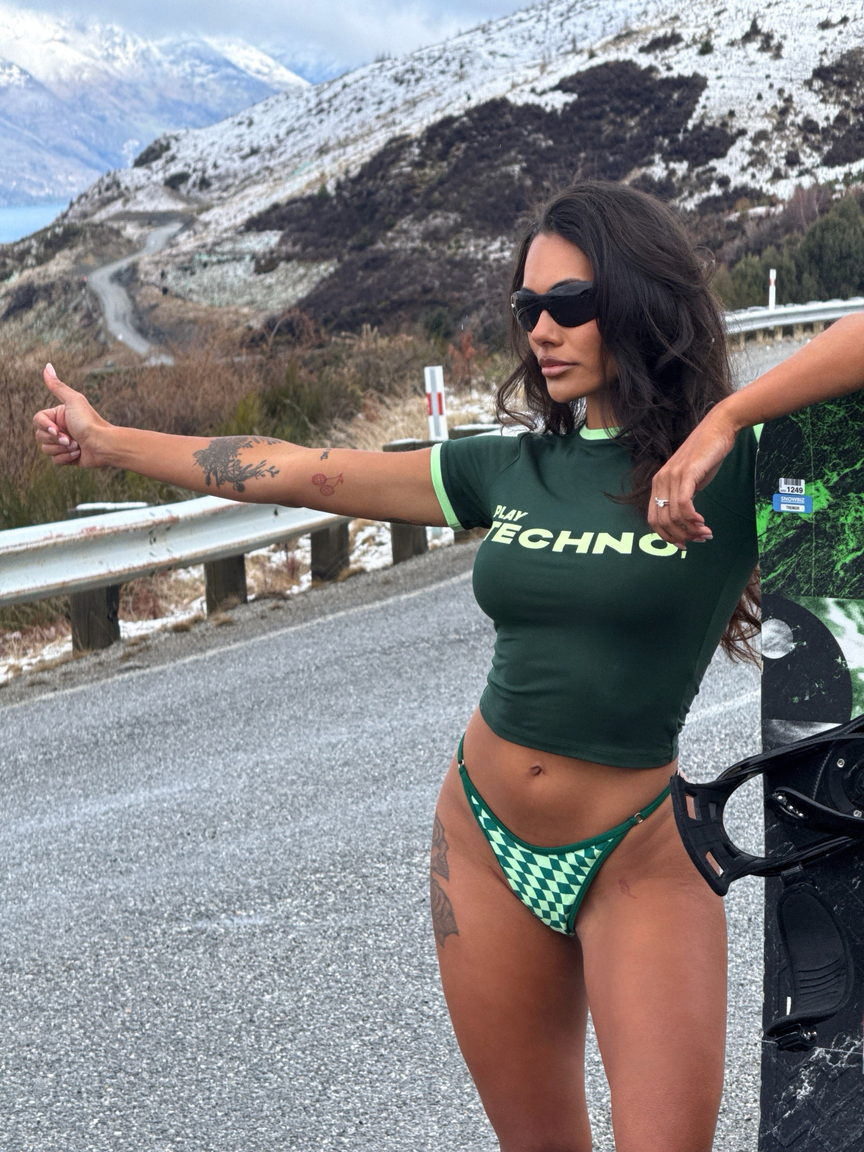 Model wearing black sunglasses, a green Rogue Season cropped t-shirt with printed text 'PLAY TECHNO' and green checkered bikini bottoms, standing on a road in a snowy setting.