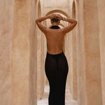 Model wearing a black halter neck dress standing in a sunlit archway with sand-coloured walls.
