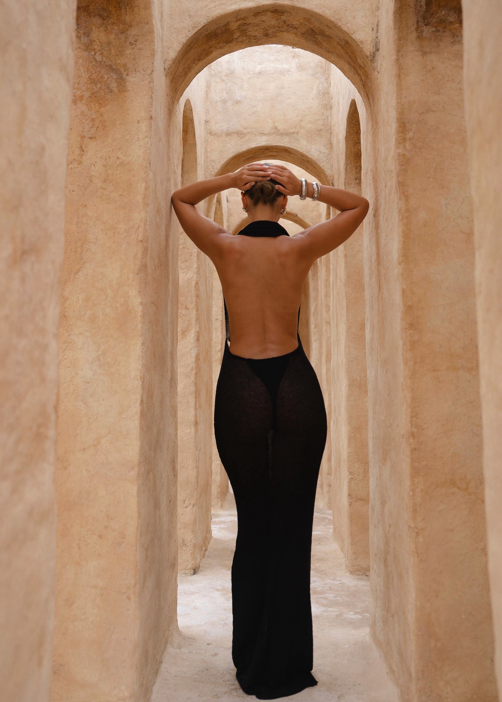 Model wearing a black halter neck dress standing in a sunlit archway with sand-coloured walls.