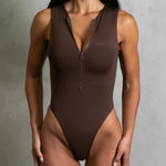Close up shot of model in a brown zip up swimsuit standing in front of a grey wall.