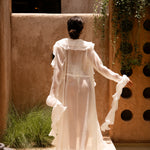 Back shot of model wearing a ivory bikini with a sheer ruffled maxi cover-up, standing outdoors in sunlight against a textured beige wall with greenery.