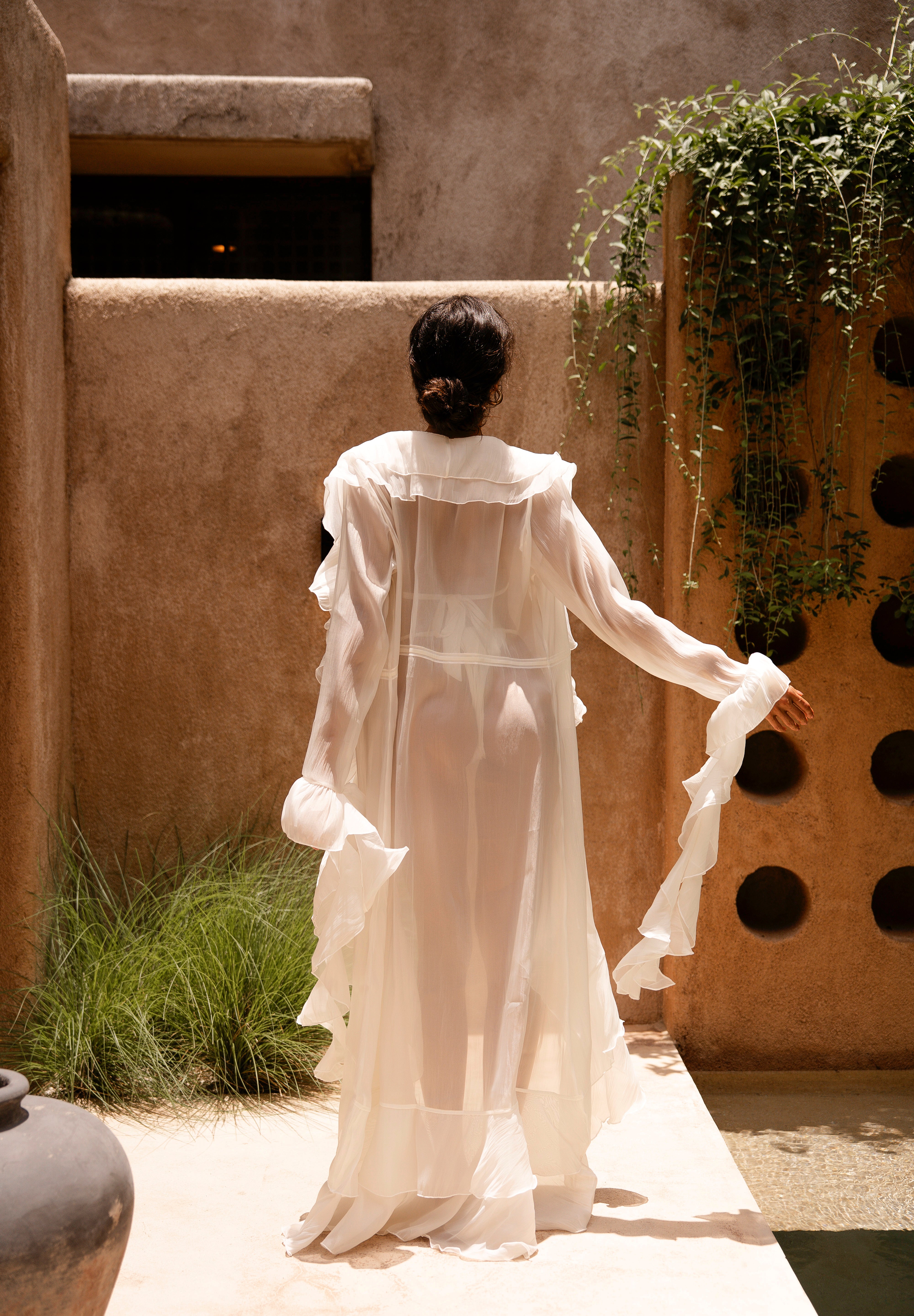 Back shot of model wearing a ivory bikini with a sheer ruffled maxi cover-up, standing outdoors in sunlight against a textured beige wall with greenery.