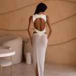 Back shot of model wearing an ivory bodysuit with a matching sheer maxi skirt with a fitted silhouette, standing indoors against a neutral-toned wall with minimal decor.