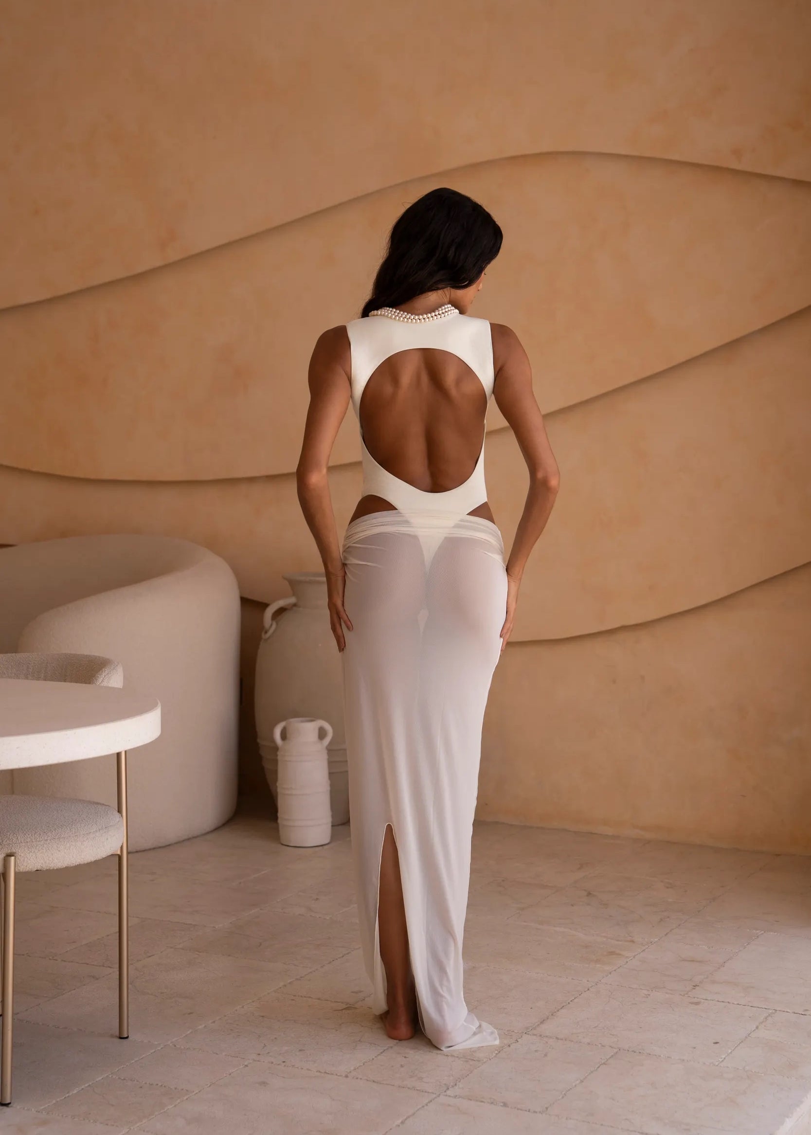 Back shot of model wearing an ivory bodysuit with a matching sheer maxi skirt with a fitted silhouette, standing indoors against a neutral-toned wall with minimal decor.