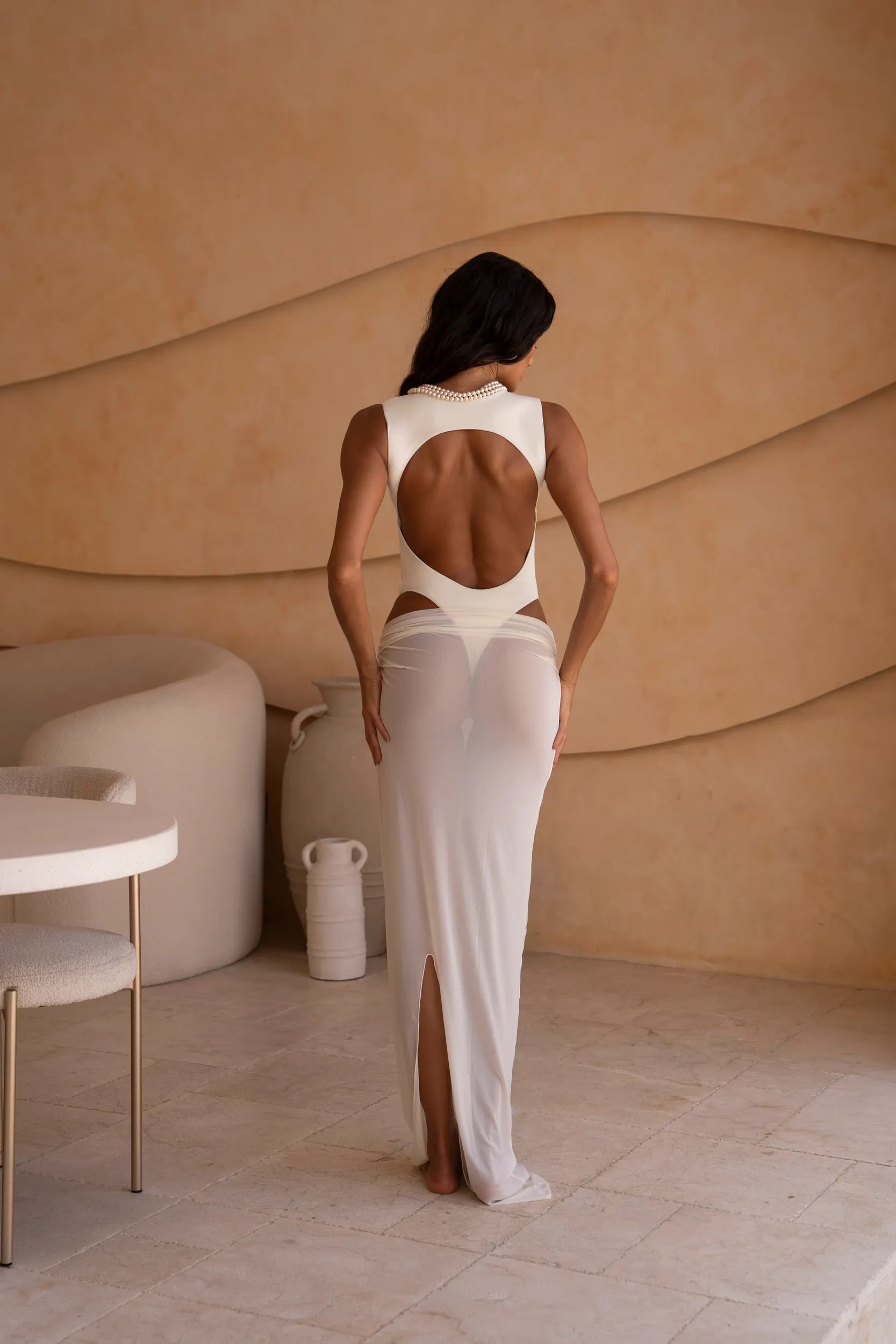 Back shot of model wearing an ivory bodysuit with a matching sheer maxi skirt with a fitted silhouette, standing indoors against a neutral-toned wall with minimal decor.