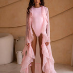 Model wearing a coral one-piece swimsuit layered with a coral sheer flowing ruffle cover-up. The look features a high-cut silhouette, soft feminine tones, and statement earrings. Set against a warm textured backdrop, the styling evokes an elegant resort and beach-club aesthetic.