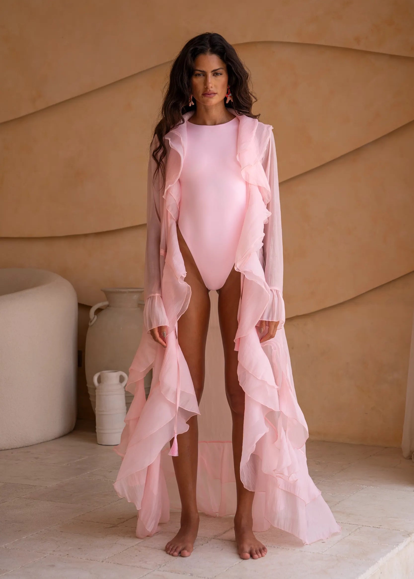 Model wearing a coral one-piece swimsuit layered with a coral sheer flowing ruffle cover-up. The look features a high-cut silhouette, soft feminine tones, and statement earrings. Set against a warm textured backdrop, the styling evokes an elegant resort and beach-club aesthetic.