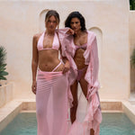 Two models pose in coordinated blush pink resortwear looks against a warm-toned textured backdrop. One model wears a coral print bikini top with matching tie-side bottoms, layered with a sheer ruffle cover-up, while the other wears a soft pink halter bikini paired with a matching sheer maxi sarong. The styling creates a feminine, elevated vacation-inspired aesthetic.