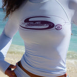 Close up shot of model wearing a baby blue long sleeve rash with a purple Rogue Season logo and matching bikini bottoms, standing on a tropical beach.