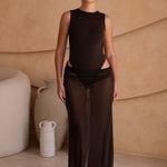 Model wearing a dark brown bodysuit with a matching sheer maxi skirt with a fitted silhouette, standing indoors against a neutral-toned wall with minimal decor.