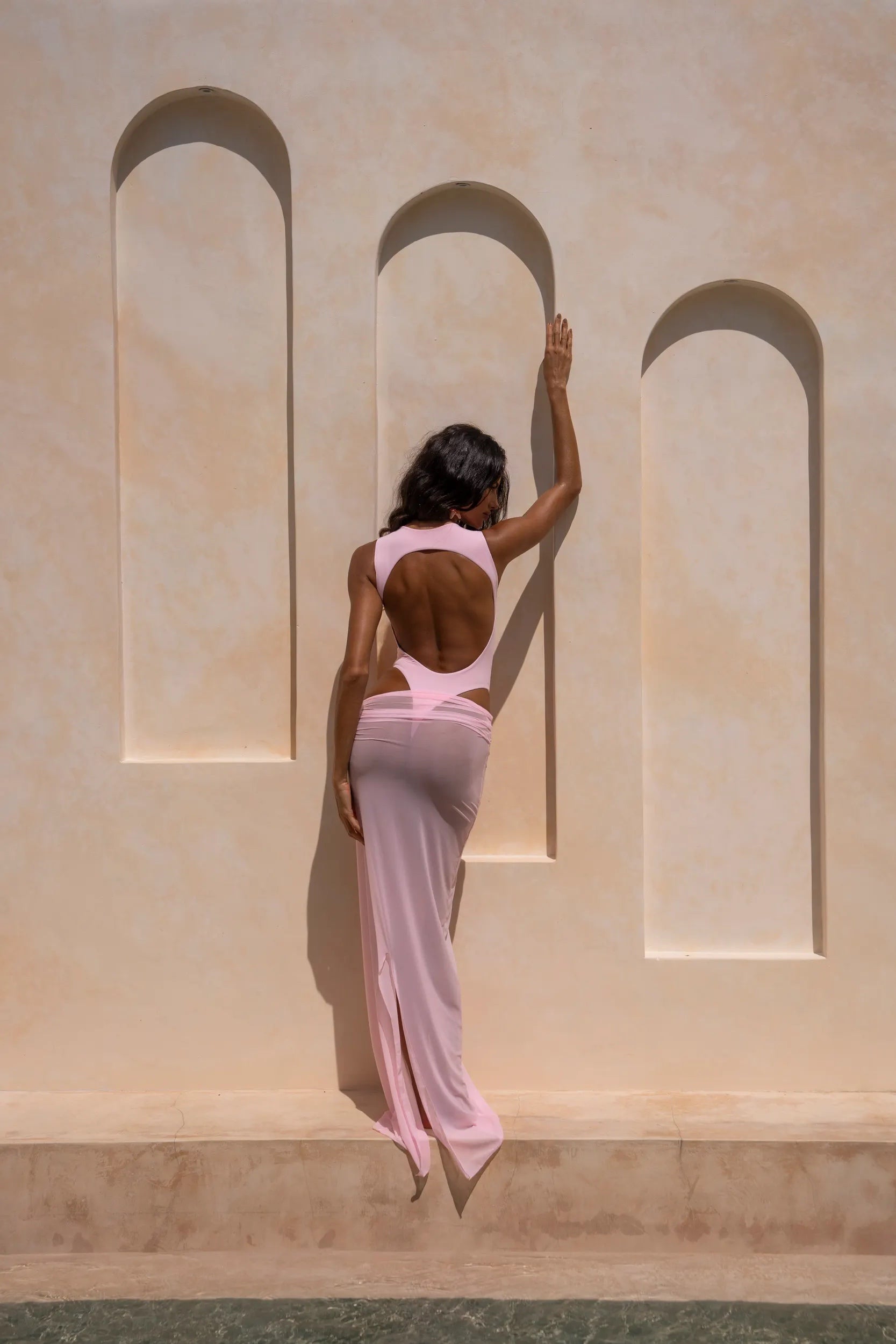 Back shot of model wearing a soft pink one-piece swimsuit with a sheer maxi skirt.