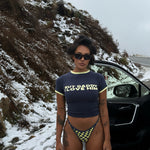 Model wearing a navy Rogue Season cropped t-shirt with printed text and navy and yellow checkered bikini bottoms, standing next to a car on a snowy road.