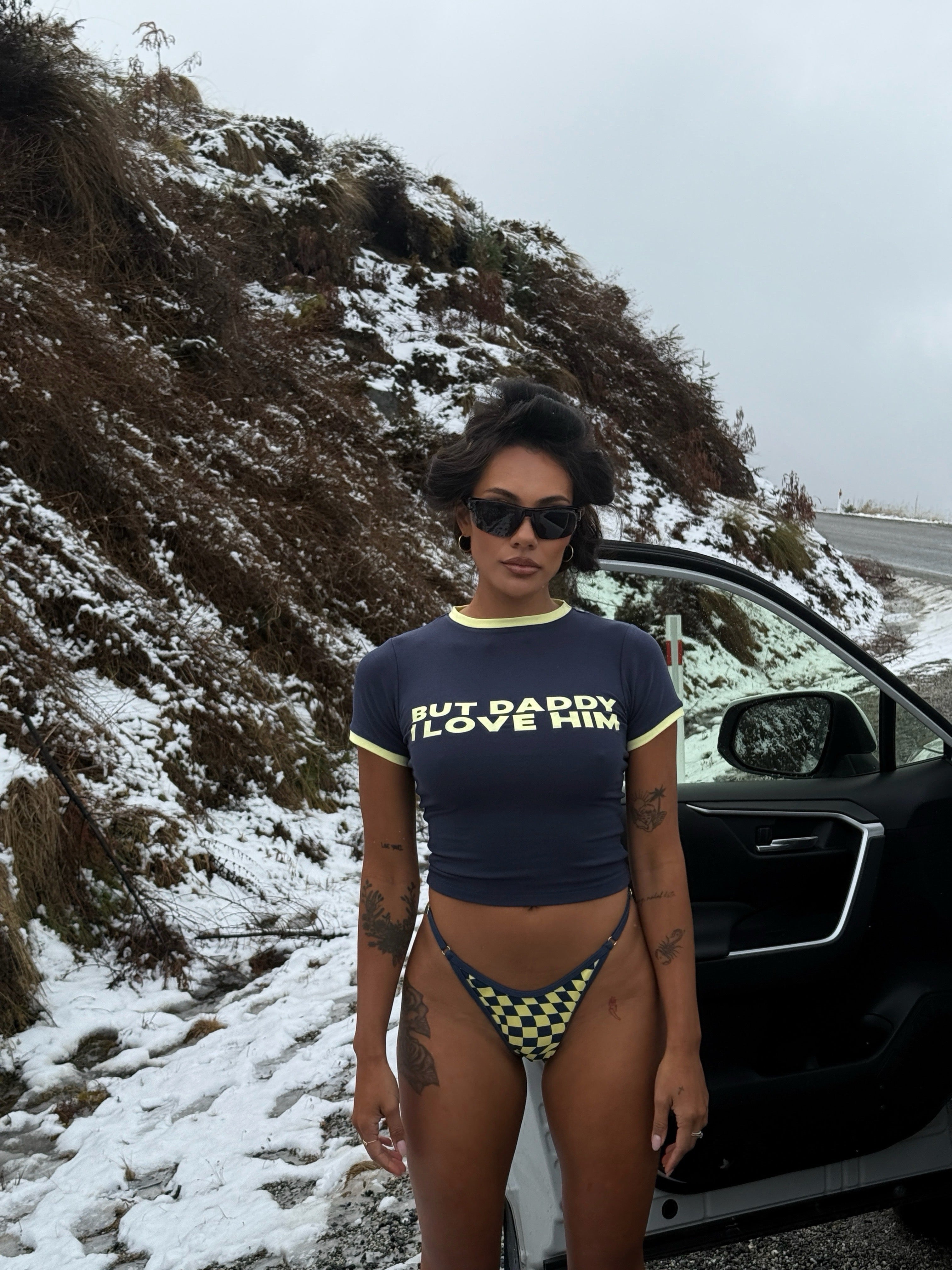 Model wearing a navy Rogue Season cropped t-shirt with printed text and navy and yellow checkered bikini bottoms, standing next to a car on a snowy road.