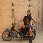 Model sitting on a motorcycle against a textured wall. She wears a brown sarong as a head scarf and she's styled it back with a black leather belt bag and black leather knee high boots.