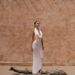 Model in a white maxi halter dress standing next to a large piece of driftwood against a beige wall.