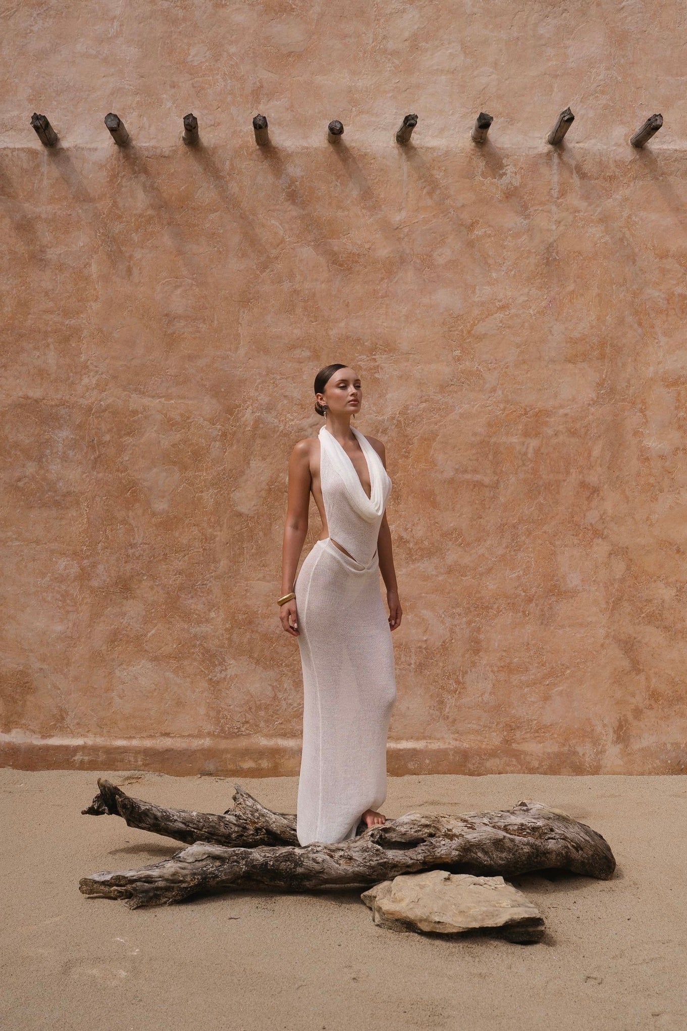 Model in a white maxi halter dress standing next to a large piece of driftwood against a beige wall.