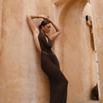 Model wearing a brown halter neck maxi dress standing in a sunlit archway with sand-coloured walls.