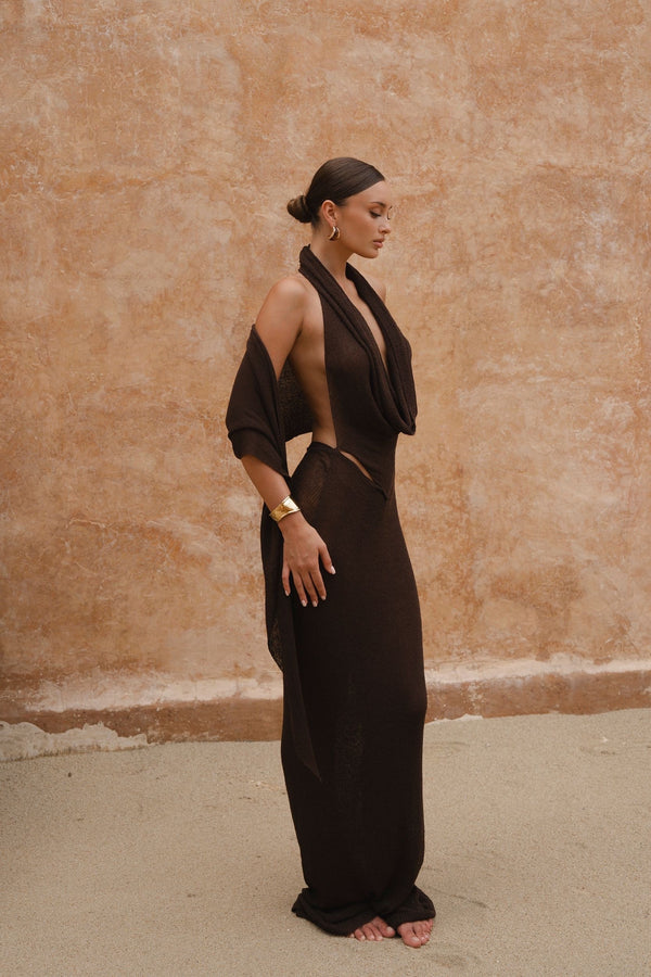 Model wearing a chocolate brown sheer maxi dress with a draped halter neckline, standing side-on against a textured neutral wall.
