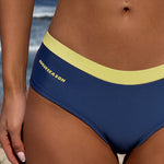 Close-up of a model wearing navy blue bikini bottoms with a light yellow waistband and yellow 'ROGUESEASON' branding on the side, set against a beach background.