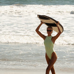 Model in a light green swimsuit holding a surfboard on a beach with ocean waves in the background