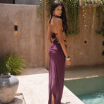 Back shot of model wearing a burgundy triangle bikini set with strappy bottoms and a matching sheer maxi skirt, standing poolside against a textured wall with greenery.