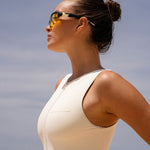 Close up side shot of model wearing an Ivory zip up swimsuit and sunglasses standing on a beach with ocean waves in the background
